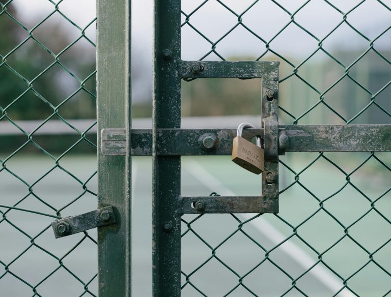 State-Owned Enterprises & Public Sector Close-up of a locked gate with chain link fence at a tennis court outdoors.
