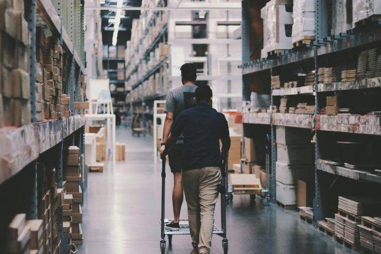 State-Owned Enterprises & Public Sector Two men maneuver a trolley in a large warehouse filled with boxes and shelves.